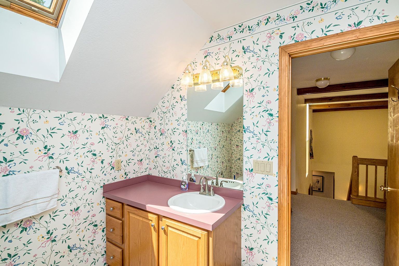 This bathroom features a vanity with a light wood cabinet and a solid-colored countertop, complemented by floral wallpaper that adds a traditional aesthetic. A large mirror sits above the sink, illuminated by a multi-bulb light fixture, while a skylight provides natural overhead lighting. The room connects directly to an adjacent carpeted hallway, offering a view of a wooden staircase.
