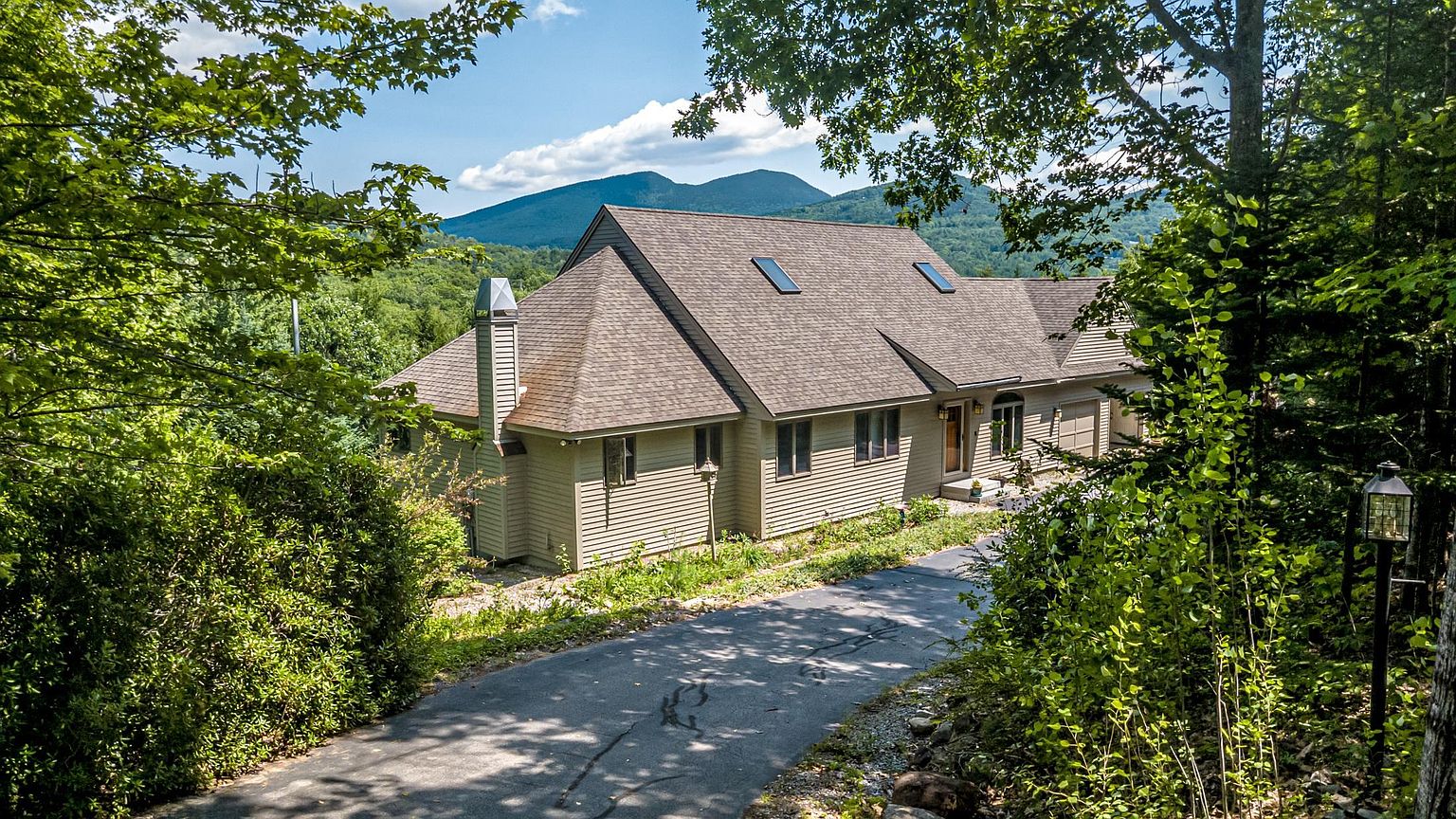 This elevated perspective captures a single-story home nestled in a lush, wooded setting with a scenic mountain backdrop. The house features light-colored horizontal siding, a multi-gabled roof with skylights, and an attached garage, all accessed by a paved driveway that winds through the trees. The surrounding mature forest and clear blue sky create a serene, private atmosphere typical of a mountain retreat.