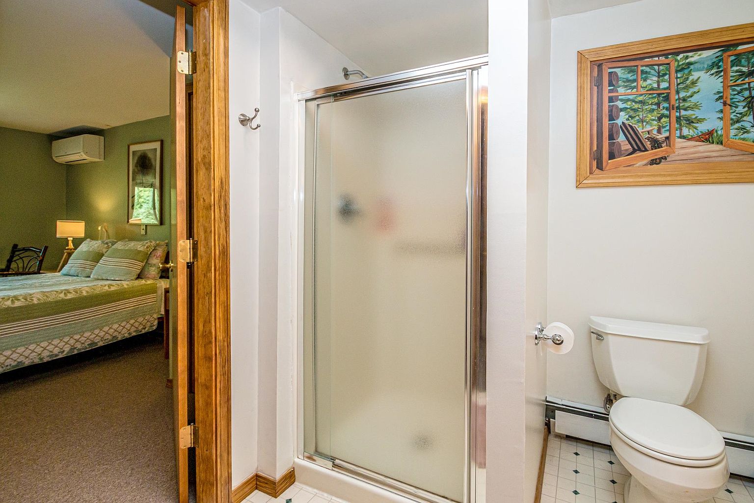 This bathroom features a glass-enclosed shower with frosted panels and a standard white toilet positioned next to a wall-mounted toilet paper holder. A framed nature-themed print hangs on the wall above the toilet, while a doorway provides a direct view into an adjacent bedroom with a bed and a wall-mounted air conditioning unit. The space is functional and compact, with a neutral color palette and simple fixtures.