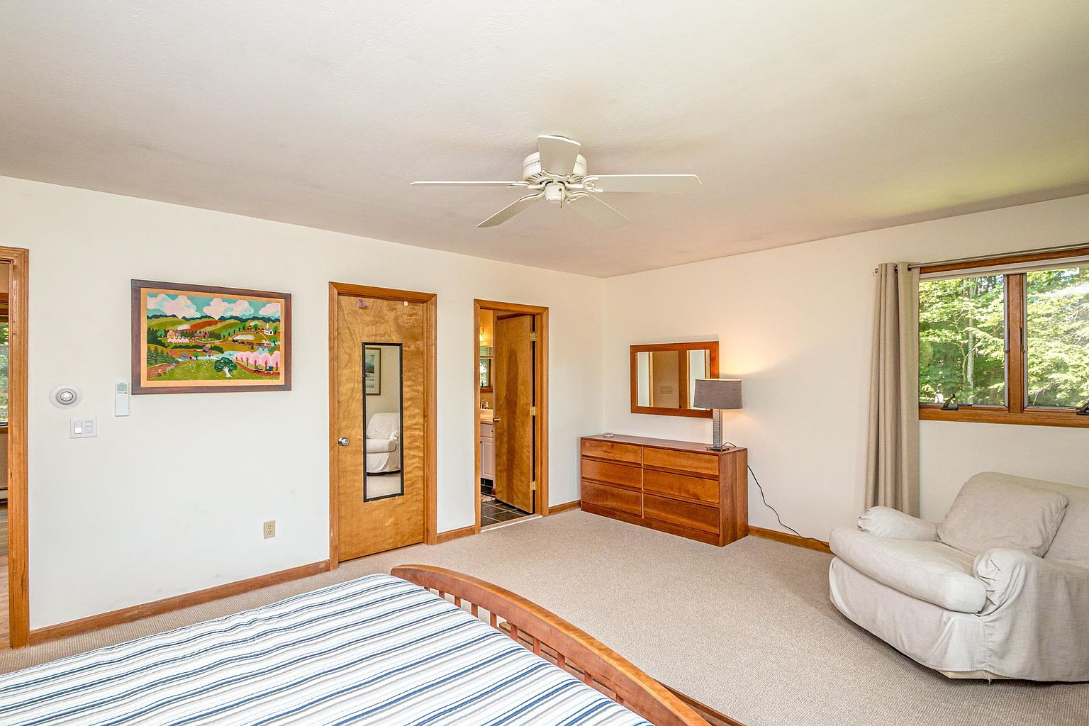 This bedroom features a neutral color palette with light-colored walls and carpet, creating a bright and airy atmosphere. The room is furnished with a wooden dresser, a matching mirror, a table lamp, and a comfortable armchair positioned near a window that looks out onto greenery. A striped bed frame is visible in the foreground, and the space includes two wooden doors leading to other areas of the home.