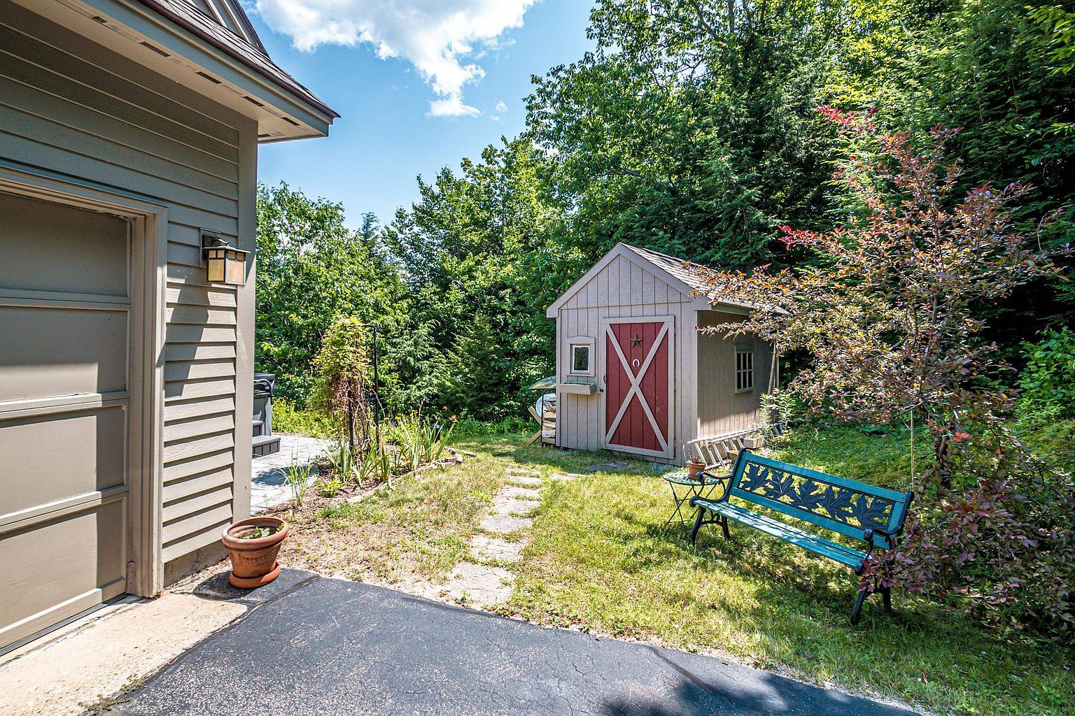 This backyard scene features a small, tan-colored storage shed with red-trimmed double doors and a decorative star accent, set against a backdrop of lush green trees. A weathered teal metal bench sits on the grass near a stone pathway that leads from the side of the garage toward the shed. The area is framed by mature landscaping, creating a quiet and private outdoor space.