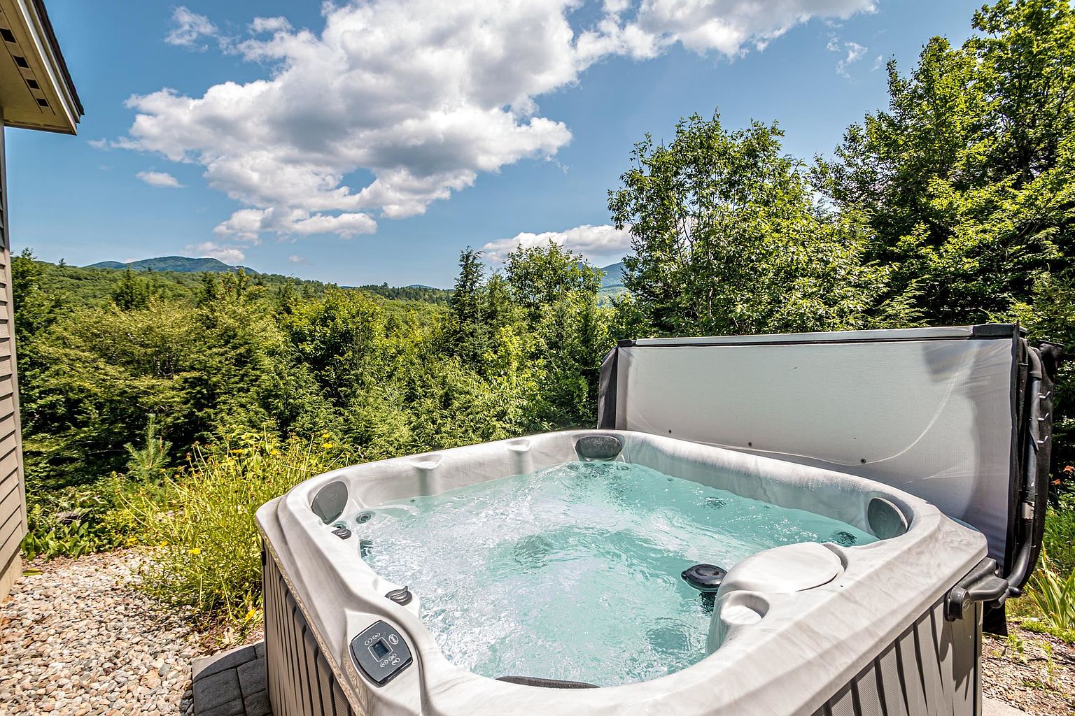 A relaxing outdoor hot tub is positioned on a stone patio, offering a panoramic view of lush green mountains and a vibrant blue sky with scattered clouds. The spa features a light-colored basin with integrated jets and a partially raised cover, set against a backdrop of dense forest foliage. This serene setting highlights the property's connection to nature and provides an inviting space for outdoor leisure.