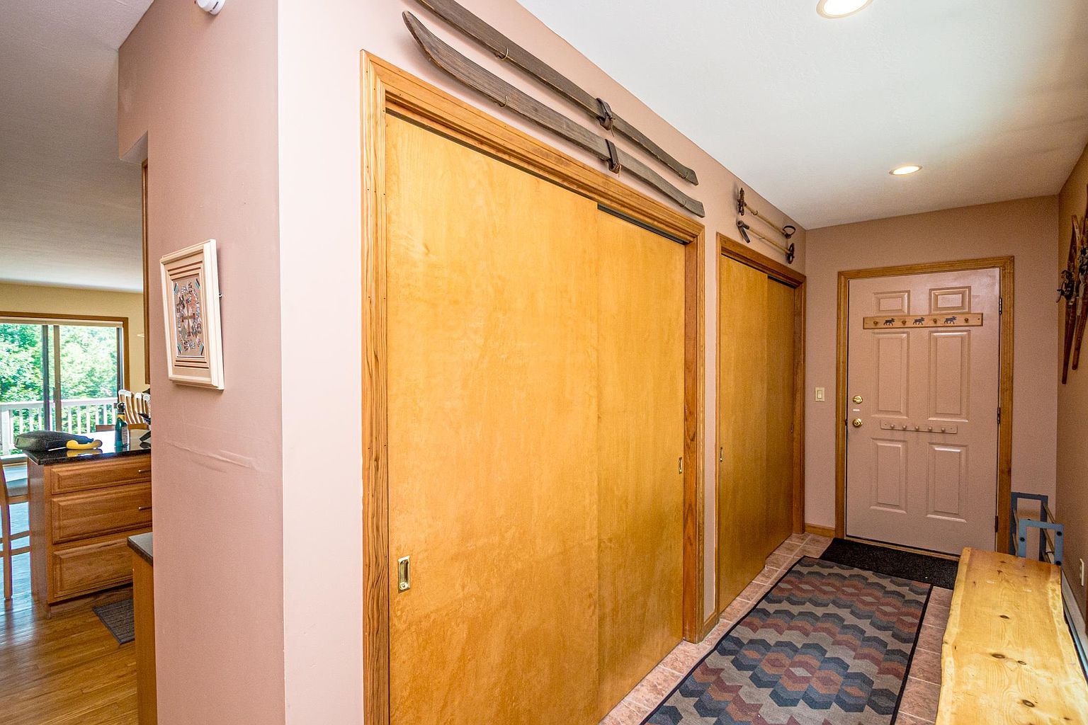 This entryway hallway features light wood-toned sliding closet doors and a patterned area rug that adds texture to the space. The walls are decorated with vintage-style wooden skis and poles, contributing to a rustic, cabin-inspired aesthetic. A wooden bench sits near the front door, which is accented with a decorative coat rack, creating a welcoming and functional transition area.