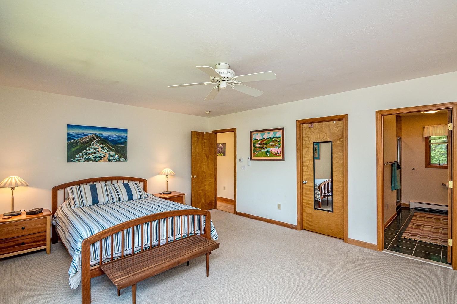 This primary bedroom features a wooden bed frame with a striped comforter, flanked by two matching nightstands with lamps. The room includes a wooden bench at the foot of the bed, two doors leading to other areas, and an open doorway revealing an attached bathroom with dark tile flooring. The space is neutrally painted with carpeted floors and a ceiling fan, offering a clean and functional layout.