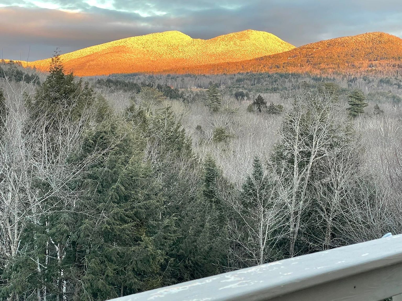This image is captured from an elevated balcony, looking out over a dense, wooded landscape toward a prominent mountain range. The setting sun casts a warm, golden glow across the mountain peaks, creating a striking contrast against the darker, shadowed forest in the foreground. The perspective emphasizes the scenic, natural views available from the property's outdoor living space.