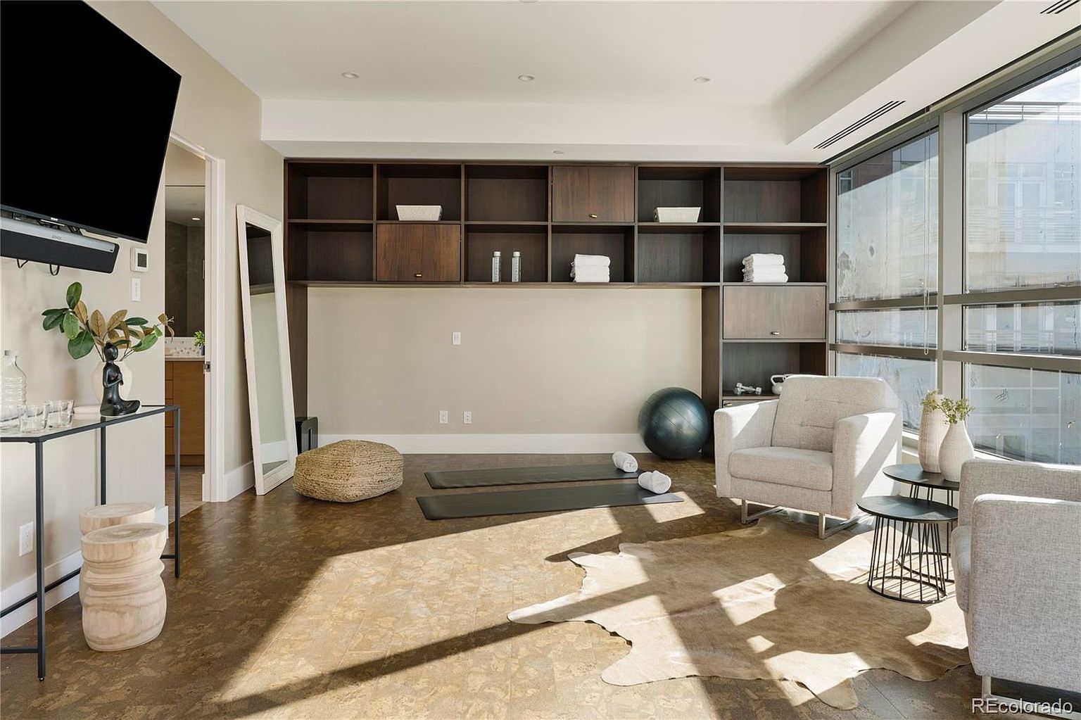 This is an interior shot of a home gym featuring a built-in shelving unit with dark wood cabinets and open shelves. The room includes workout mats, a yoga ball, and comfortable seating, suggesting a versatile space for both exercise and relaxation. Large windows provide ample natural light, enhancing the airy and inviting atmosphere.
