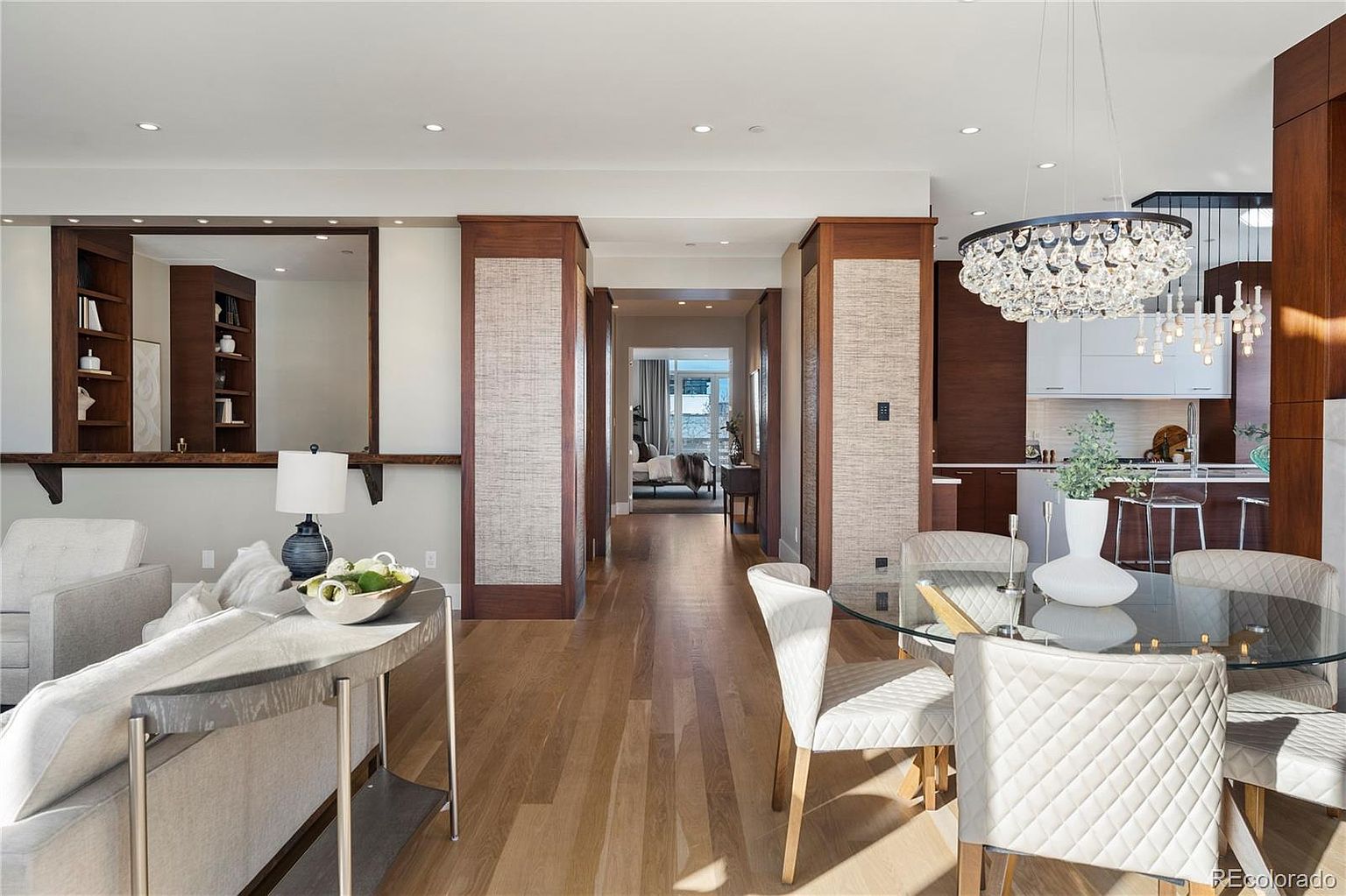 This interior shot showcases a dining area adjacent to a living space and kitchen. The dining area features a glass-topped round table surrounded by white upholstered chairs with a crystal chandelier hanging above. The space is defined by wooden pillars with textured panels, leading to a hallway, and the hardwood flooring adds warmth to the room.