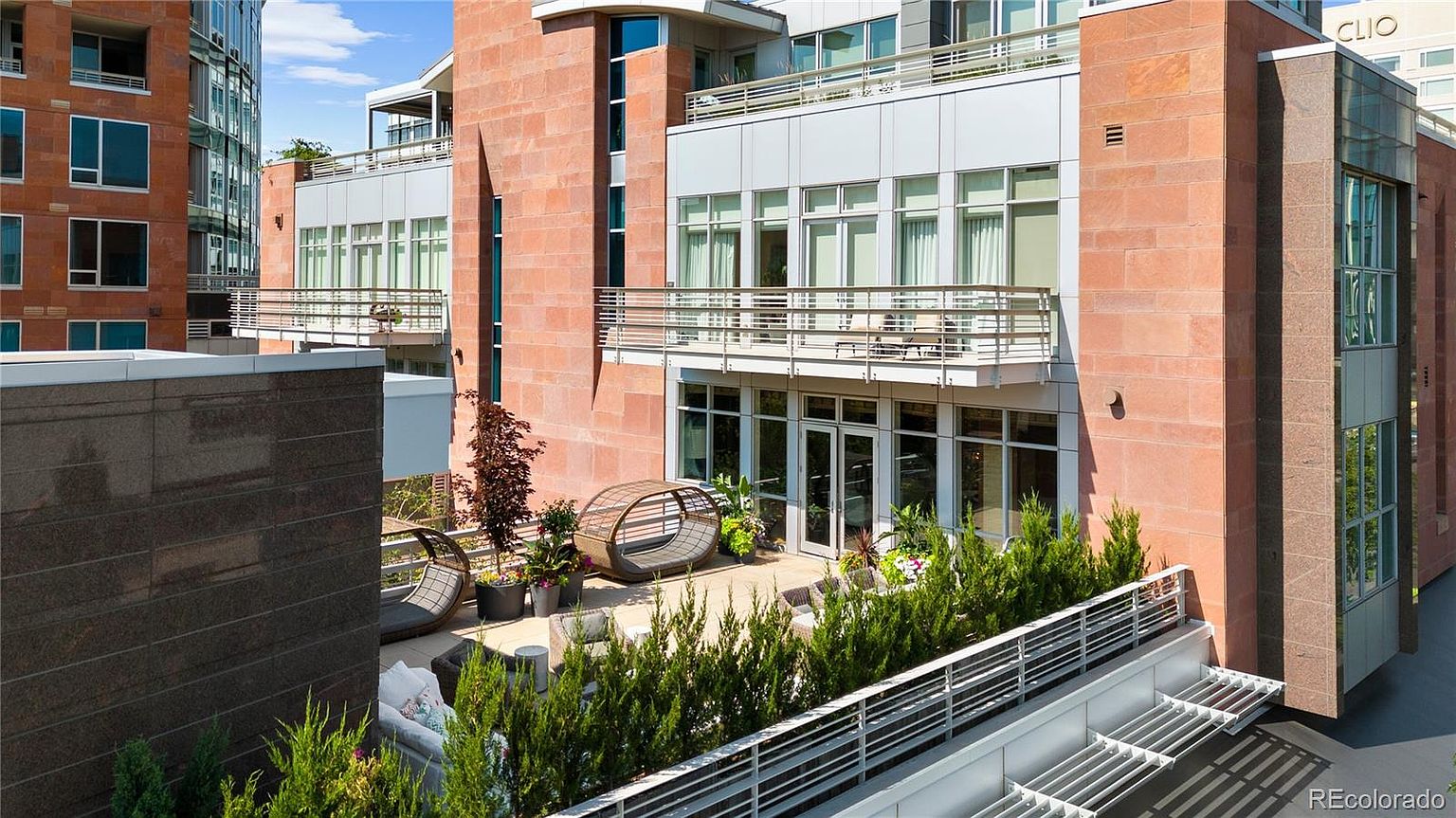 This image showcases a luxurious patio/deck area attached to a modern building. The space is furnished with stylish outdoor seating, including a unique circular lounger and a comfortable sofa, all surrounded by lush greenery. The patio is elevated, offering a private outdoor retreat with a contemporary design.