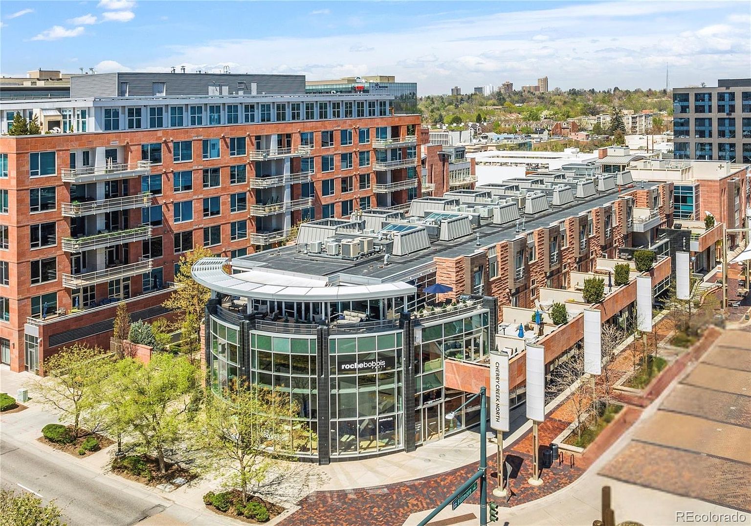 This aerial view showcases a mixed-use development featuring brick buildings with modern architectural elements. The scene includes a curved glass-fronted structure, likely a commercial space, and residential buildings with balconies. The surrounding landscape includes trees and a glimpse of the cityscape in the background, creating an urban yet inviting atmosphere.