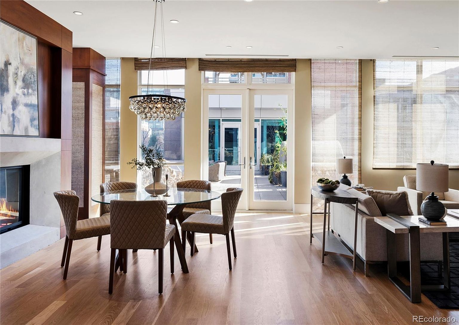 This is an interior shot of a dining room featuring a round glass table with upholstered chairs. A modern chandelier hangs above the table, and large windows with woven shades provide natural light and a view of the outdoor patio. The room has hardwood floors and a fireplace, creating a warm and inviting atmosphere.