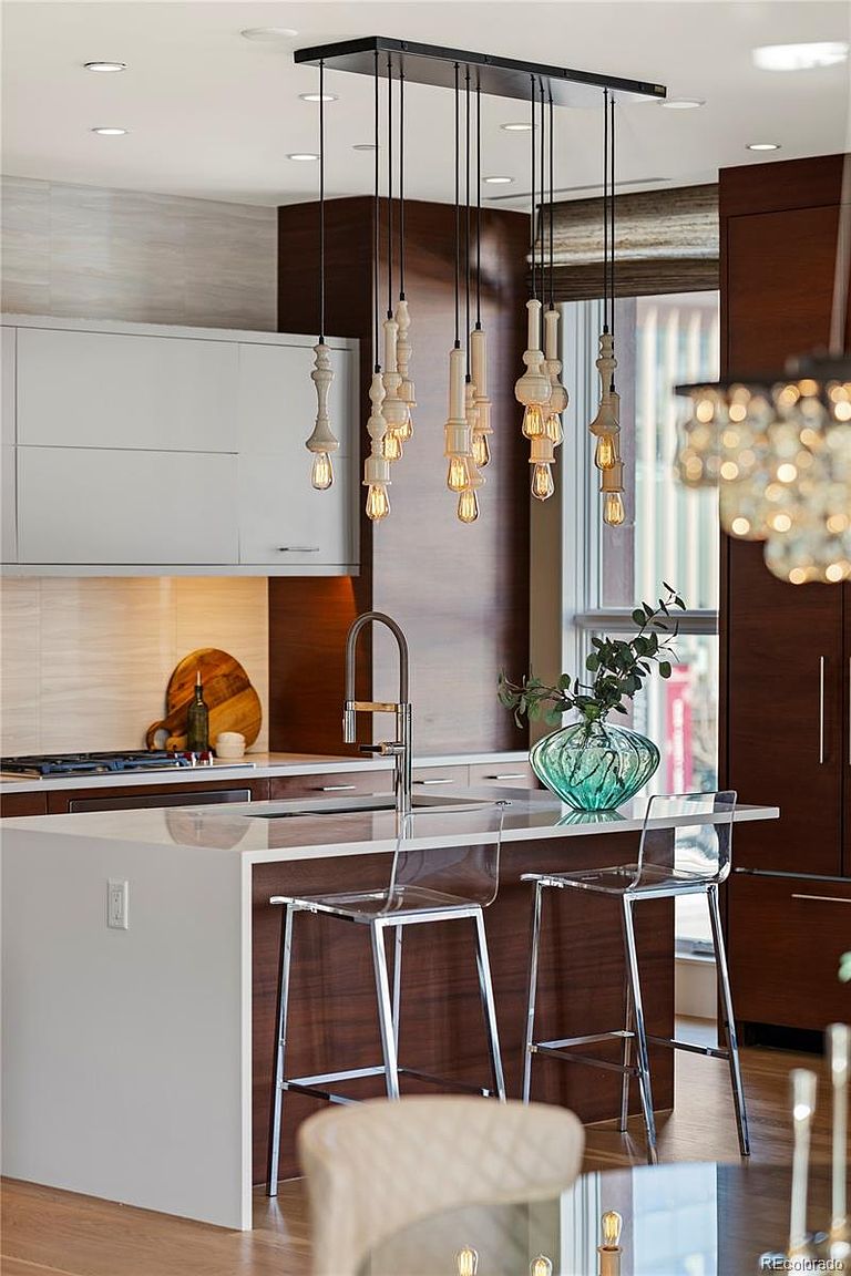 This is a well-lit kitchen featuring a modern design with a white countertop island and dark wood cabinetry. The island has clear bar stools and a decorative vase with greenery. A unique pendant lighting fixture hangs above the island, adding a touch of elegance and style to the space.