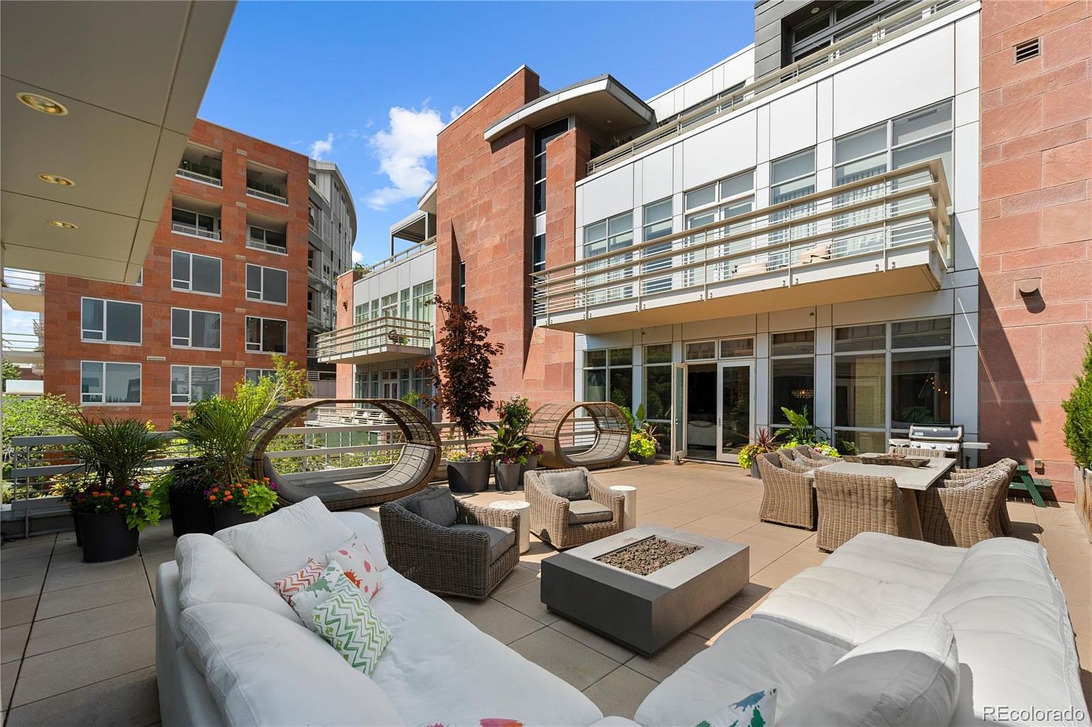 This image showcases a luxurious outdoor patio or balcony area, furnished with comfortable white sofas, wicker chairs, and a modern fire pit. The space is designed for relaxation and entertainment, with additional seating options and potted plants adding to the ambiance. The surrounding architecture features a mix of brick and modern white facades, suggesting a high-end residential building.