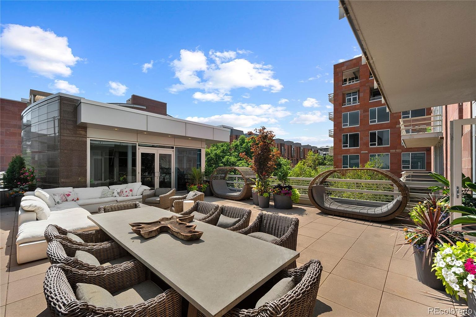 This image showcases a luxurious outdoor patio or balcony area, furnished with a large dining table surrounded by wicker chairs, a comfortable sectional sofa, and unique pod-like lounge chairs. The space is adorned with potted plants, creating a serene and inviting atmosphere, while the surrounding architecture suggests a high-end residential building with views of the cityscape and sky.