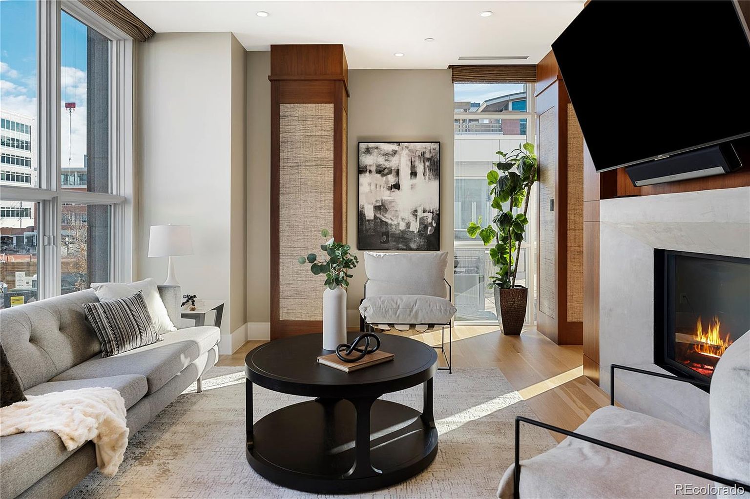 This is an interior shot of a modern living room featuring a gray sofa with decorative pillows, a round black coffee table, and a fireplace with a mounted television. Large windows provide natural light, and the room is decorated with neutral tones and contemporary art. The perspective is from a medium angle, showcasing the layout and design elements of the space.
