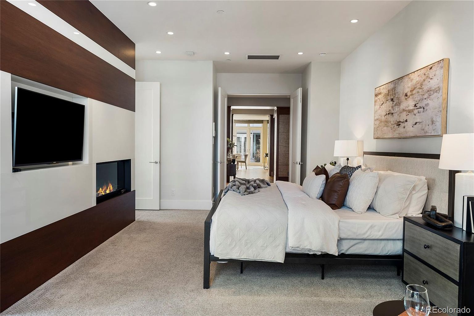 This is a primary bedroom featuring a modern design with a fireplace and TV built into a wall unit. The room has a neutral color palette with white walls and light carpet, complemented by dark wood accents. A large bed with multiple pillows and a side table with a lamp create a comfortable and inviting atmosphere.