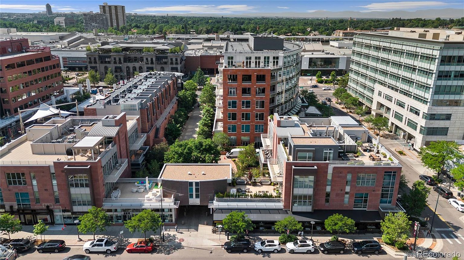 This aerial view showcases a modern urban development with a mix of residential and commercial buildings. The architecture features brick facades, rooftop terraces, and contemporary design elements. Lush greenery is interspersed throughout the area, creating a vibrant and appealing environment.