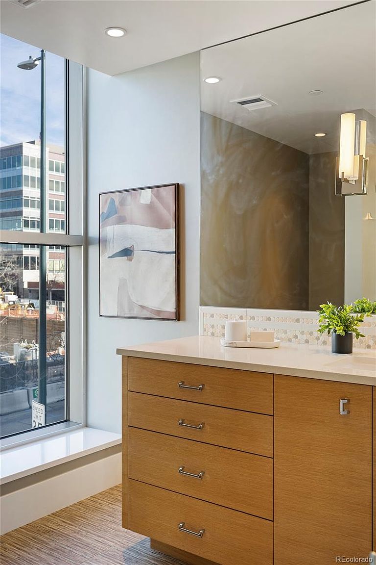 This is a well-lit primary bathroom featuring a modern vanity with light wood cabinetry and sleek silver hardware. A large mirror reflects the space, and a piece of abstract art hangs on the wall beside a window offering a city view. The overall impression is clean, contemporary, and bright.