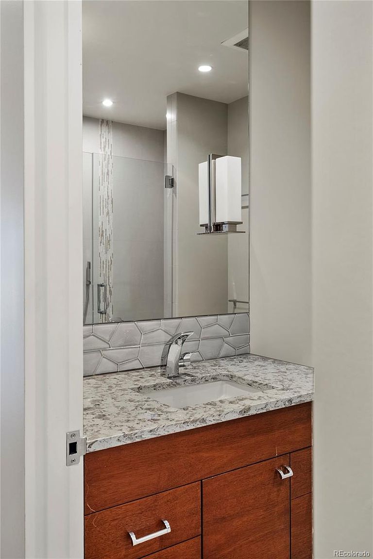 This is a bathroom featuring a vanity with a wood-finish cabinet and a light-colored granite countertop. A modern faucet and a rectangular sink are visible. Above the vanity is a large mirror reflecting a shower with a glass door and decorative tile. A modern light fixture is mounted on the wall next to the mirror. The perspective is from the doorway, offering a partial view of the bathroom.