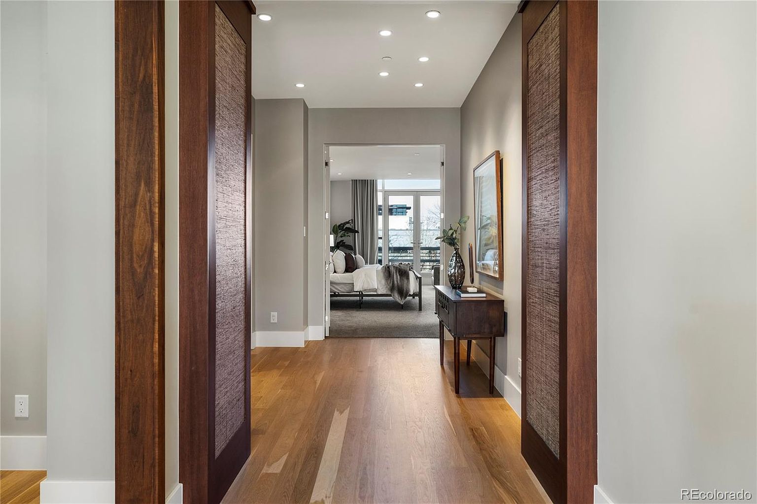 The image showcases a well-lit hallway leading into a bedroom. The hallway features hardwood flooring, light gray walls, and decorative wooden door frames with textured panels. A small console table with a vase and artwork adds a touch of elegance, while the bedroom beyond offers a glimpse of a comfortable and inviting space.