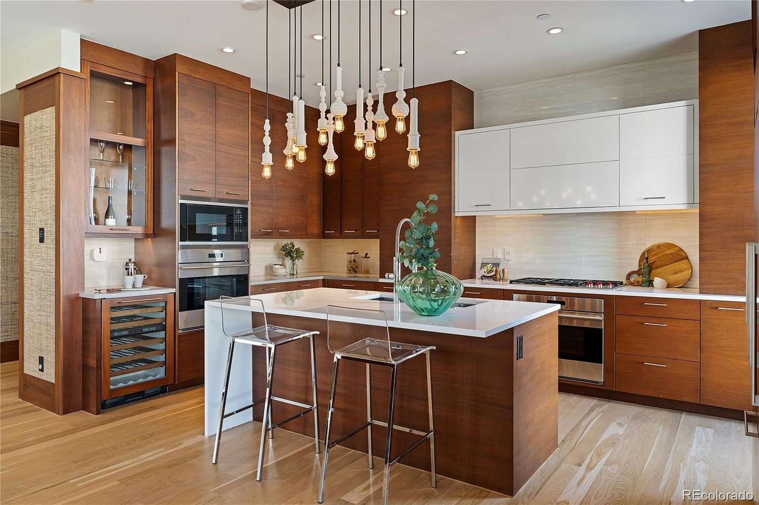 This is a well-lit, modern kitchen featuring rich wood cabinetry contrasted with white upper cabinets and a white countertop island. The kitchen is equipped with stainless steel appliances, including a built-in microwave and oven, and a wine cooler. Unique pendant lighting hangs above the island, which is complemented by clear bar stools, creating an inviting and stylish space.