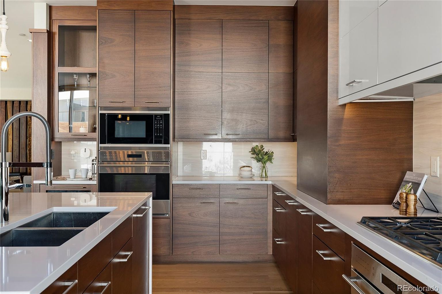 This is a modern kitchen with dark wood cabinetry and white countertops. Stainless steel appliances are built into the cabinetry, and a large island with a sink is in the foreground. The kitchen has a clean and sophisticated design.
