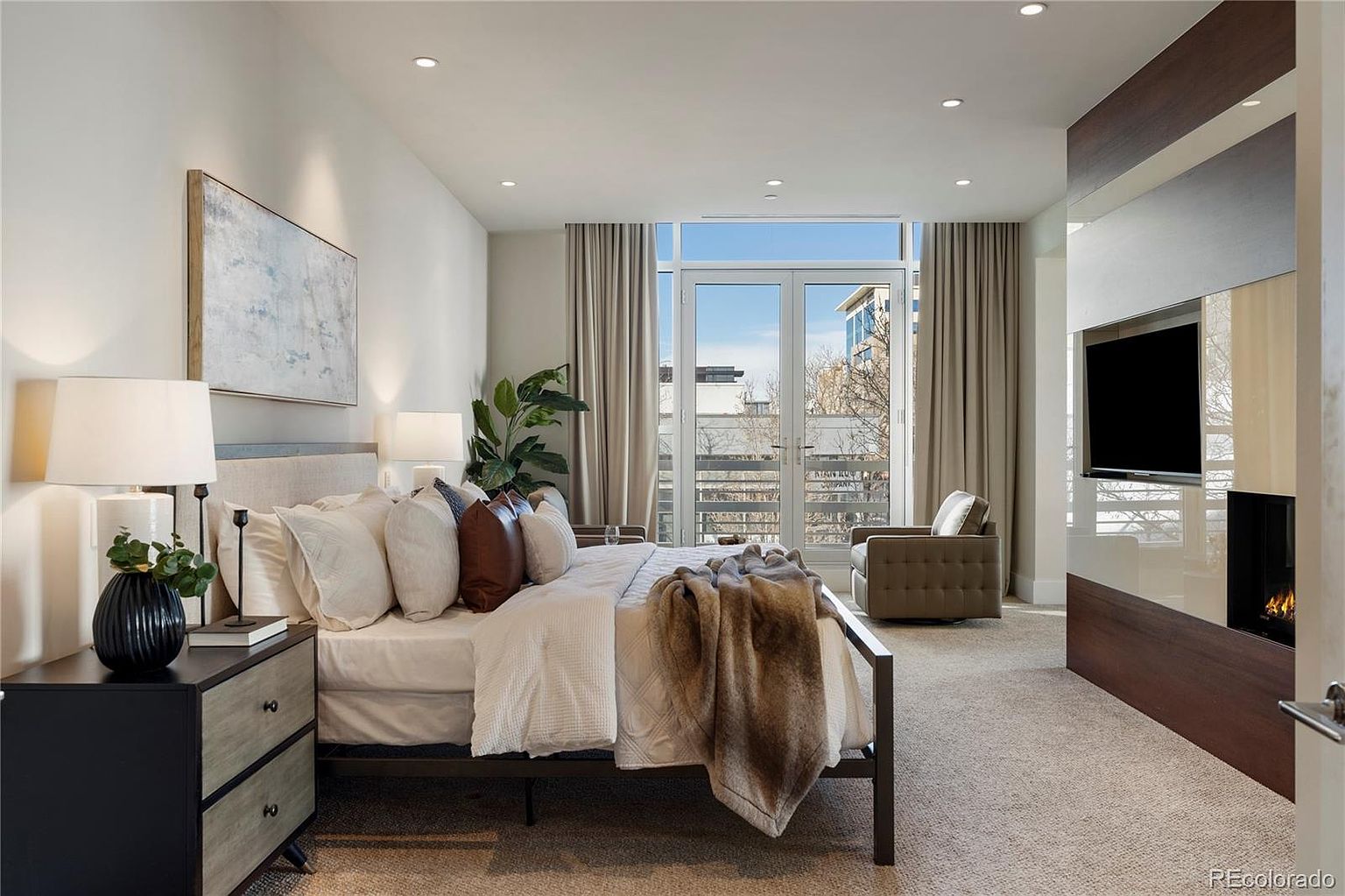 This is a primary bedroom featuring a bed with a neutral-toned headboard, decorative pillows, and a throw blanket. The room has large windows with curtains, a side table with lamps, and a modern entertainment center with a TV and fireplace. The overall style is contemporary and luxurious.