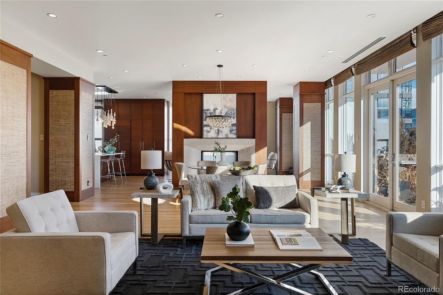 This is an interior shot of a luxurious living room. The room features a neutral color palette with hardwood floors, a dark patterned rug, and modern furniture including a sofa, armchairs, and a coffee table. A fireplace with a decorative mantle and artwork above it serves as a focal point, and large windows provide ample natural light.