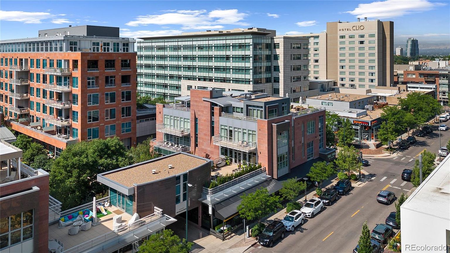 This aerial view showcases a vibrant urban landscape with a mix of modern and contemporary architecture. Several multi-story buildings with brick and glass facades dominate the scene, complemented by lush greenery and well-maintained streets lined with parked cars. The overall impression is one of a thriving, upscale urban environment.