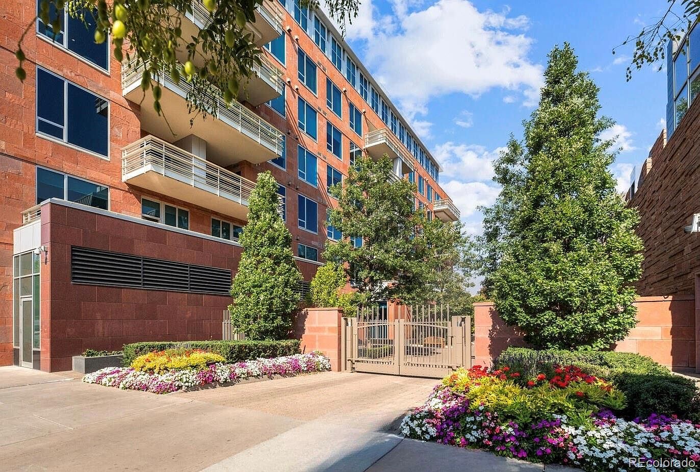 This is a front view of a modern brick apartment building with multiple floors and balconies. The entrance features a gated driveway with meticulously maintained flower beds on either side, adding a touch of elegance and curb appeal. The building's architecture is contemporary, with clean lines and large windows.