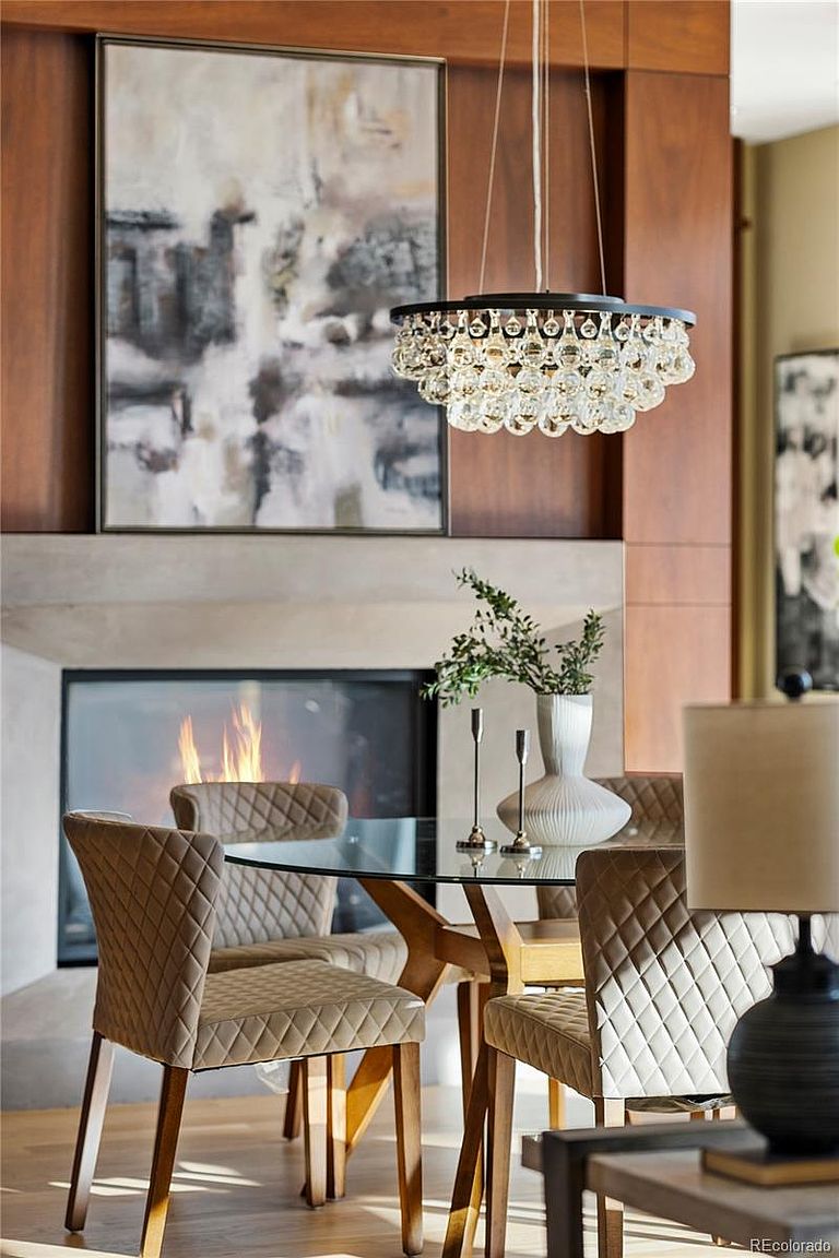 This interior shot showcases a dining room with a modern aesthetic. A round glass-top table is surrounded by upholstered chairs with a quilted pattern, set against a backdrop of a fireplace and abstract art. A crystal chandelier hangs above, adding a touch of elegance to the space.