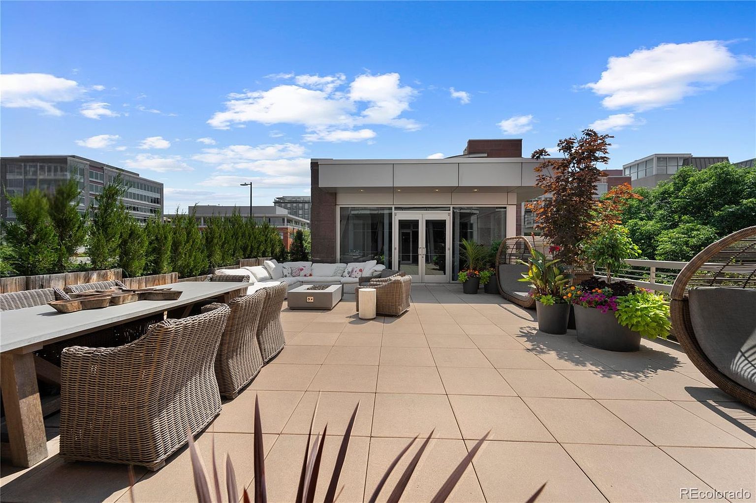 This image showcases a luxurious rooftop patio or balcony, featuring stylish outdoor furniture including a dining table with wicker chairs, a comfortable lounge area with a fire pit, and hanging chairs. The space is adorned with potted plants and trees, creating a serene and inviting atmosphere. The patio offers views of the surrounding cityscape, enhancing the appeal of this outdoor living space.