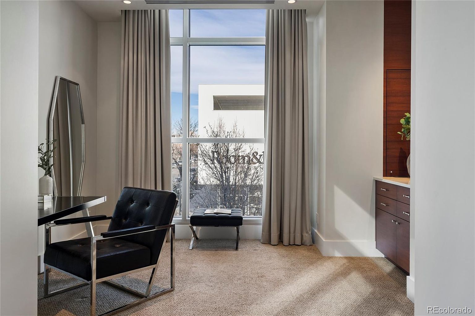 This is an interior shot of a modern office or study. The room features a black leather chair with a chrome frame, a black desk, and a matching ottoman. Large windows with neutral-toned curtains provide natural light, and a built-in cabinet with wood paneling adds a touch of sophistication.