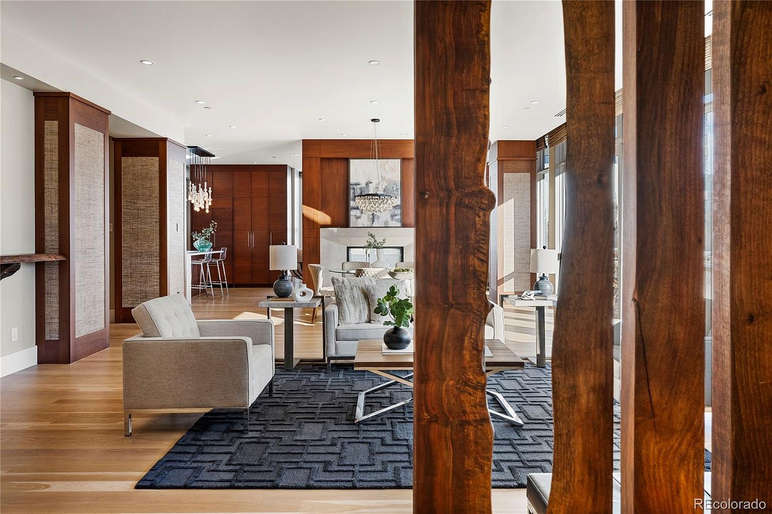 This is an interior shot of a modern living room featuring hardwood floors, a dark blue patterned rug, and contemporary furniture. The room is divided by wooden slat partitions, adding a unique architectural element. The space is well-lit with recessed lighting and natural light, creating a warm and inviting atmosphere.