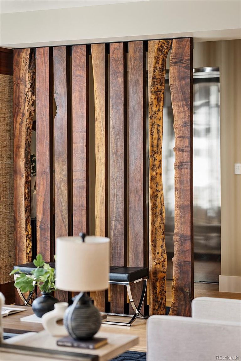 This interior shot showcases a stylish living room featuring a unique wooden slat room divider with natural wood edges, adding a rustic yet modern touch. The foreground includes a coffee table with decorative items and a lamp, while a glimpse of a seating area and a hallway can be seen in the background. The overall impression is one of sophisticated design and comfortable living.
