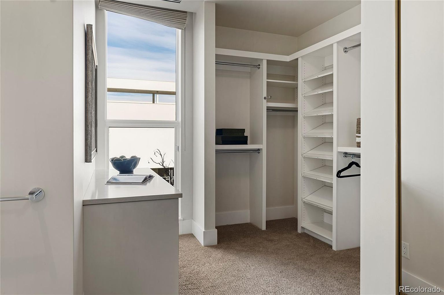 This is a well-organized walk-in closet featuring custom white shelving and hanging rods. The closet is brightly lit, possibly by natural light from a nearby window, and has neutral-toned carpeting. The design maximizes storage space and presents a clean, modern aesthetic.