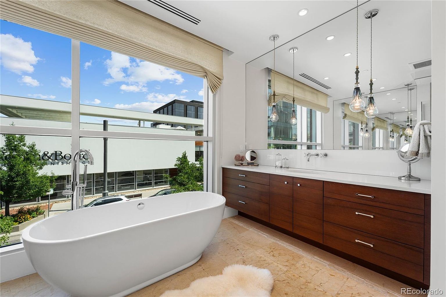 This is a luxurious primary bathroom featuring a freestanding white bathtub positioned in front of a large window with a view of the city. The bathroom boasts a long, dark wood vanity with a white countertop and a large mirror with pendant lighting. The flooring is a light-colored tile, and a plush rug adds a touch of comfort.