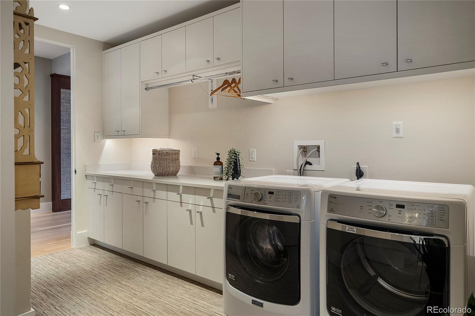 This is a well-organized laundry room featuring white cabinetry above and below a countertop, providing ample storage space. A hanging rod is installed beneath the upper cabinets for drying clothes. The room includes a modern washer and dryer set, and the flooring appears to be a neutral-toned rug or carpet, creating a clean and functional space.
