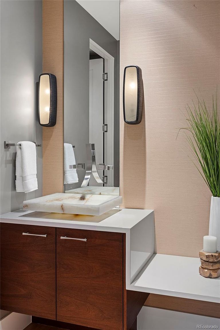 This is a modern bathroom featuring a unique marble sink atop a dark wood vanity with white countertop extension. The walls are a combination of gray and textured beige, complemented by contemporary sconce lighting and a large mirror. A decorative plant adds a touch of nature to the space.