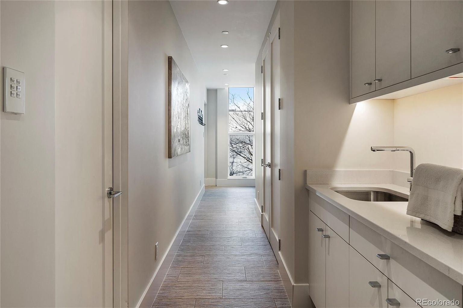 This is an interior shot of a hallway featuring light gray walls and wood-look tile flooring. The hallway is well-lit with recessed lighting and natural light coming from a window at the end of the hall. On the right, there is a utility sink and cabinets, suggesting a laundry area or butler's pantry.