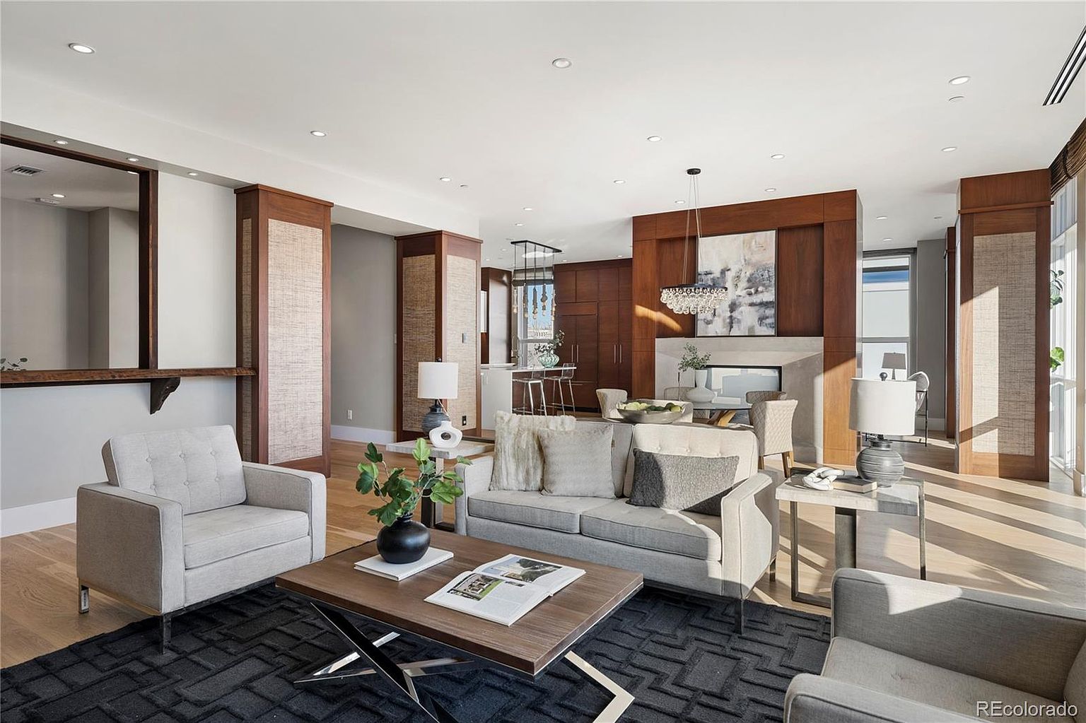 This is an interior shot of a modern living room featuring a neutral color palette with wood accents. The room is furnished with a sofa, armchairs, a coffee table, and side tables, all arranged on a dark patterned rug. A fireplace and decorative lighting add to the ambiance, creating a warm and inviting space.