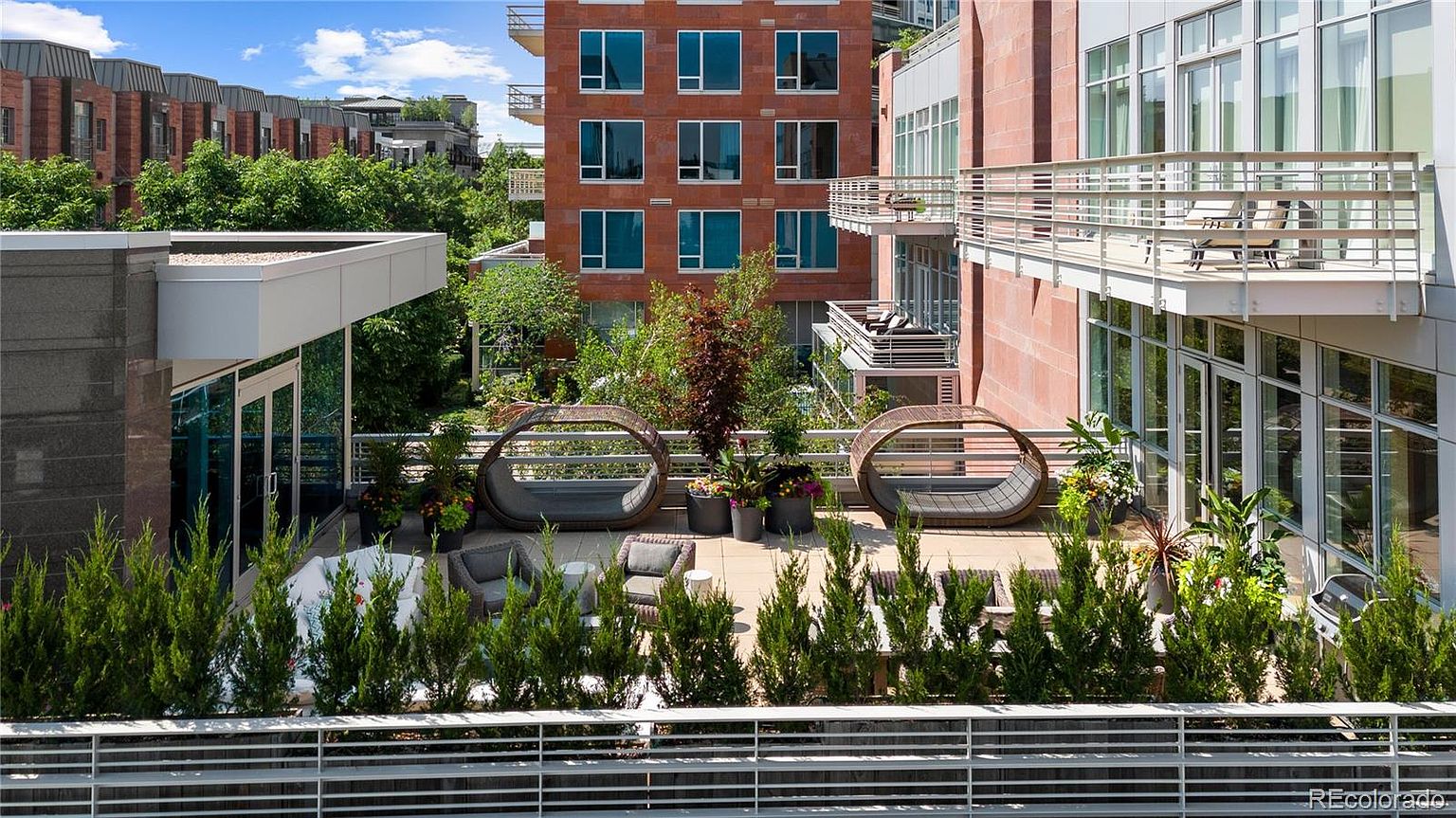 This image showcases a luxurious outdoor patio or balcony area, featuring modern lounge seating and lush greenery. The space is well-appointed with stylish furniture, including circular daybeds and comfortable chairs, creating an inviting atmosphere for relaxation and entertainment. The surrounding architecture includes brick buildings and ample windows, suggesting an upscale urban setting.