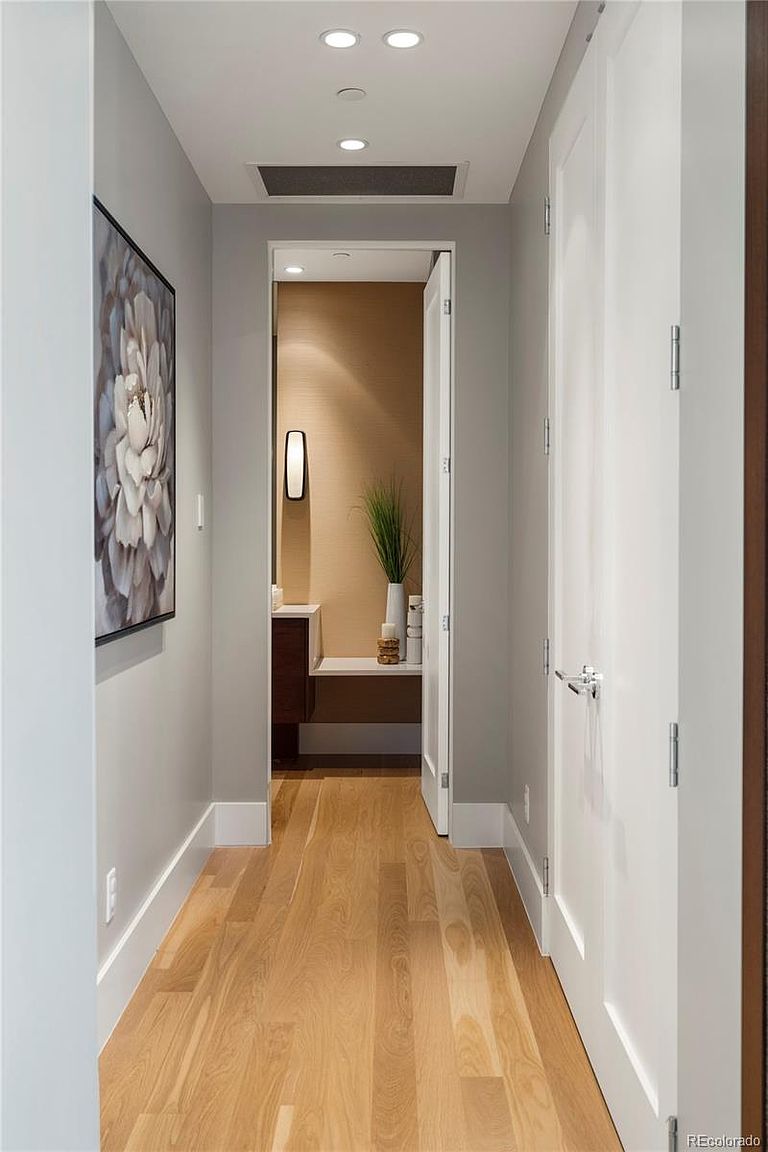 This is an interior shot of a hallway featuring light hardwood floors and light gray walls. A framed floral artwork hangs on the left wall, and a partially open white door leads to a bathroom with a modern vanity and wall sconce. The hallway is well-lit with recessed lighting, creating a clean and inviting atmosphere.