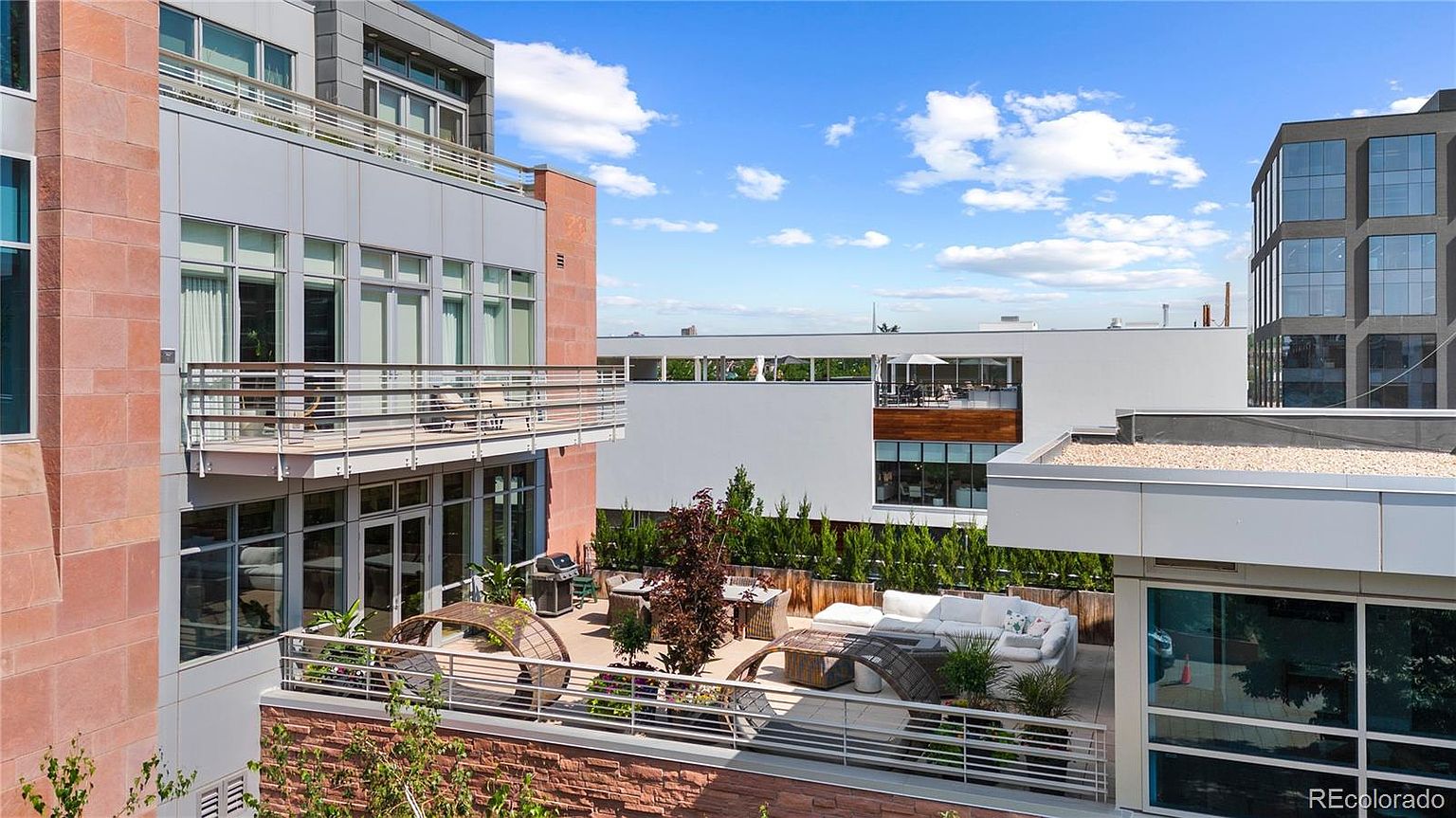 This image showcases a luxurious outdoor patio/deck/balcony area of a modern building. The space is furnished with comfortable seating, including a white sofa and wicker chairs, and features a grill and dining table, suggesting an ideal space for entertaining. The patio is surrounded by greenery, providing privacy and a touch of nature, while the overall design exudes sophistication and relaxation.