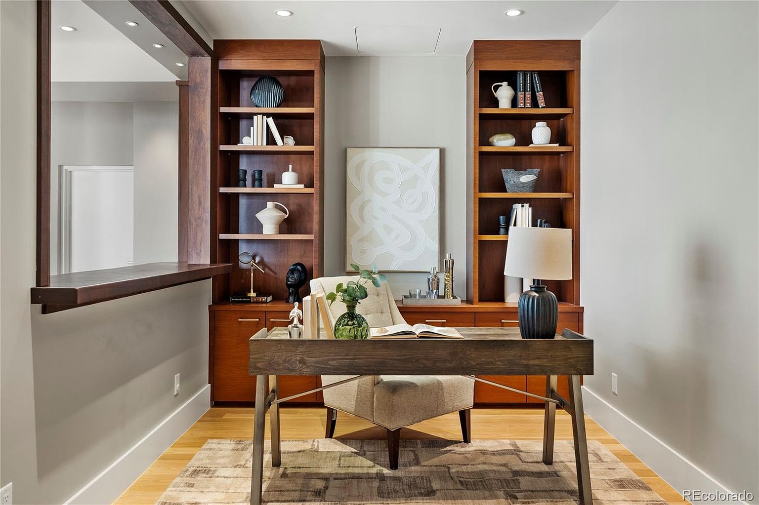 This is an interior shot of a well-appointed home office. The room features built-in wooden bookshelves flanking a central artwork, a wooden desk with metal legs, and a comfortable chair. The space is well-lit and appears to be designed for productivity and comfort, with a neutral color palette and stylish decor.