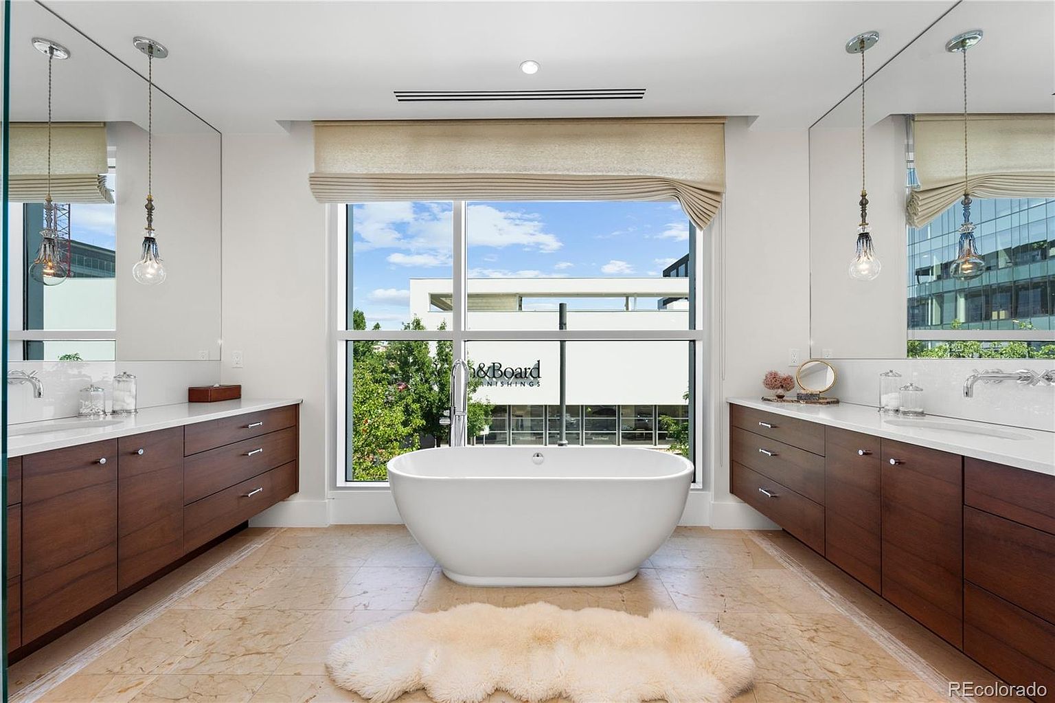 This is a luxurious primary bathroom featuring a freestanding soaking tub positioned in front of a large window with a view of the outdoors. The room is designed with dual vanities featuring dark wood cabinetry and white countertops, complemented by modern pendant lighting and a neutral color palette. A plush rug adds a touch of comfort to the marble tile flooring.