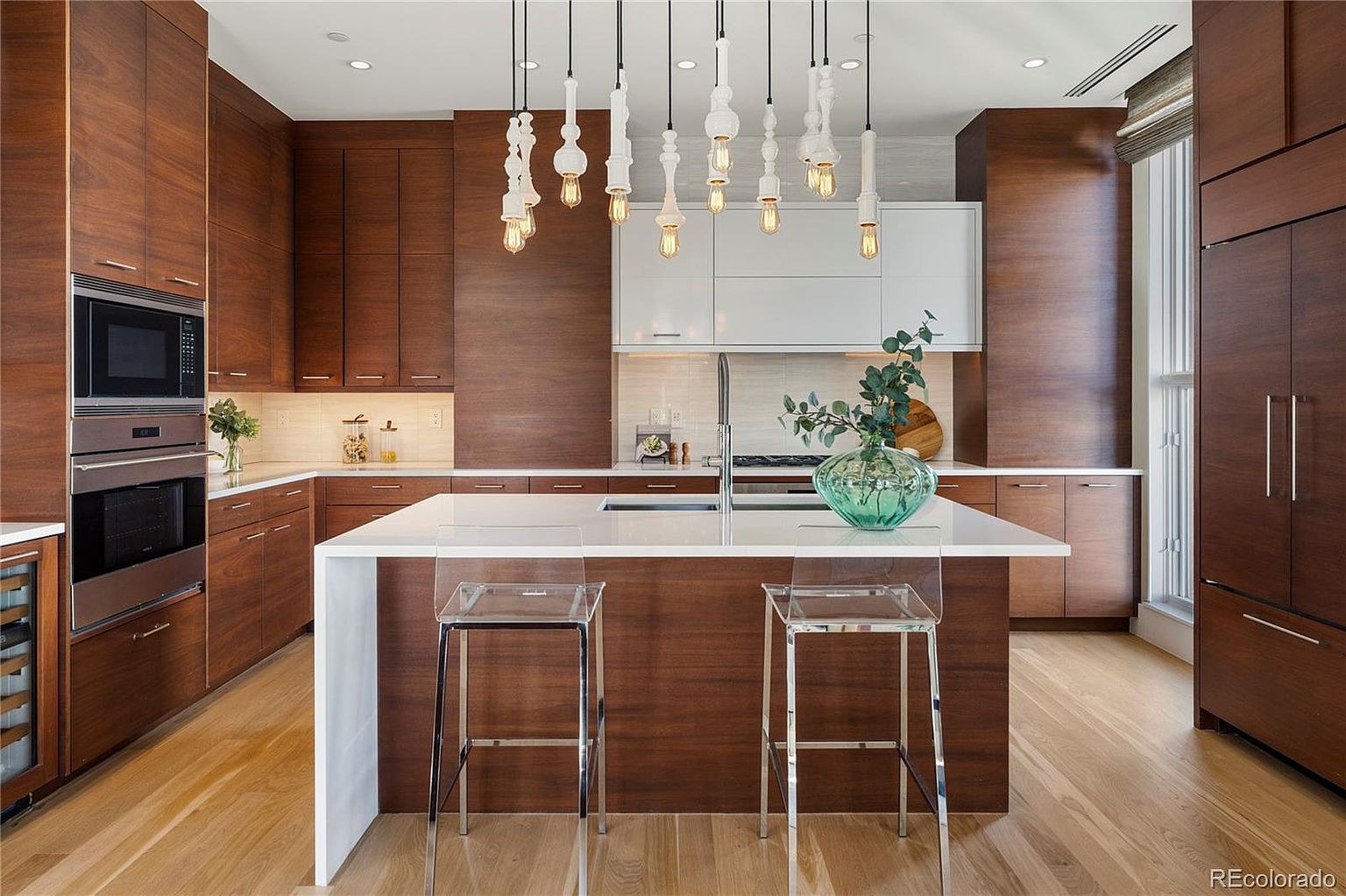 This is a modern kitchen featuring sleek, dark wood cabinetry contrasted by a white island and upper cabinets. The kitchen is illuminated by unique pendant lighting fixtures hanging above the island, which is complemented by clear bar stools. Stainless steel appliances are integrated into the cabinetry, and a large window provides natural light, creating a sophisticated and functional space.