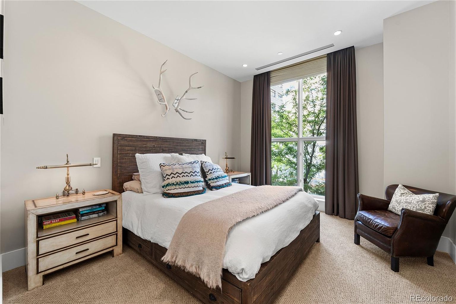 This is a cozy bedroom featuring a wooden bed frame with a white comforter and decorative pillows. A rustic nightstand with a lamp sits beside the bed, and a leather armchair is positioned near the window with brown curtains. The room has a warm and inviting atmosphere, perfect for a guest room.