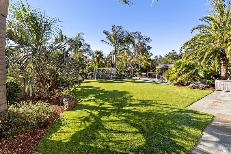 This image showcases a lush, well-maintained backyard featuring a vibrant green lawn, mature palm trees, and a soccer goal. A covered outdoor structure is visible in the background, suggesting an entertainment area. The overall impression is one of a private and inviting outdoor space perfect for recreation and relaxation.