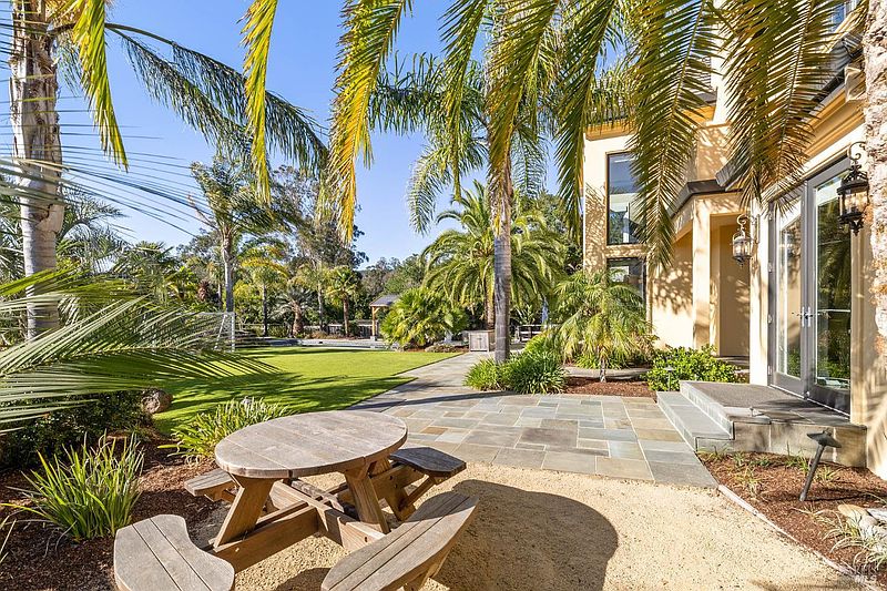 This image showcases a beautifully landscaped backyard featuring lush green grass, mature palm trees, and a charming wooden picnic table set on a gravel area. A stone pathway leads to the house, which has a light-colored exterior and glass doors, creating an inviting outdoor living space perfect for relaxation and entertaining.