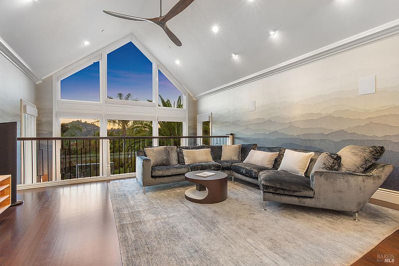 This is a spacious living room featuring a large sectional sofa, a round coffee table, and a striking wall mural depicting a mountain range. The room has high ceilings with a large A-frame window offering natural light and views of the outdoors. The overall impression is luxurious and comfortable.